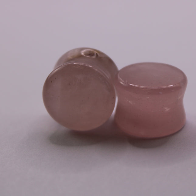 A beautifully polished pair of Rose Quartz Double Flare Plugs rests delicately on a pale surface, their soft pink tones glowing in natural light. Each plug features the gentle, translucent hue that rose quartz is known for—ranging from milky blush to faint petal pink, with subtle natural marbling throughout. The smooth, glossy finish highlights the stone’s calming energy, while the double flare design ensures a secure, comfortable fit for stretched ears.