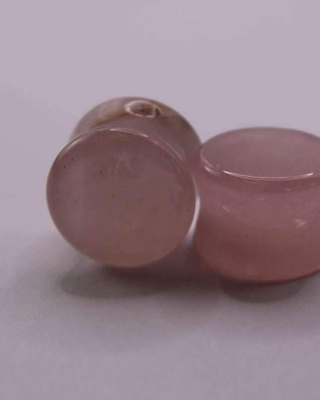 A beautifully polished pair of Rose Quartz Double Flare Plugs rests delicately on a pale surface, their soft pink tones glowing in natural light. Each plug features the gentle, translucent hue that rose quartz is known for—ranging from milky blush to faint petal pink, with subtle natural marbling throughout. The smooth, glossy finish highlights the stone’s calming energy, while the double flare design ensures a secure, comfortable fit for stretched ears. 