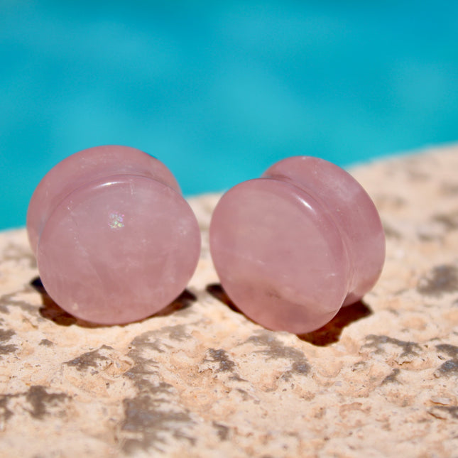 A beautifully polished pair of Rose Quartz Double Flare Plugs rests delicately on a pale surface, their soft pink tones glowing in natural light. Each plug features the gentle, translucent hue that rose quartz is known for—ranging from milky blush to faint petal pink, with subtle natural marbling throughout. The smooth, glossy finish highlights the stone’s calming energy, while the double flare design ensures a secure, comfortable fit for stretched ears.