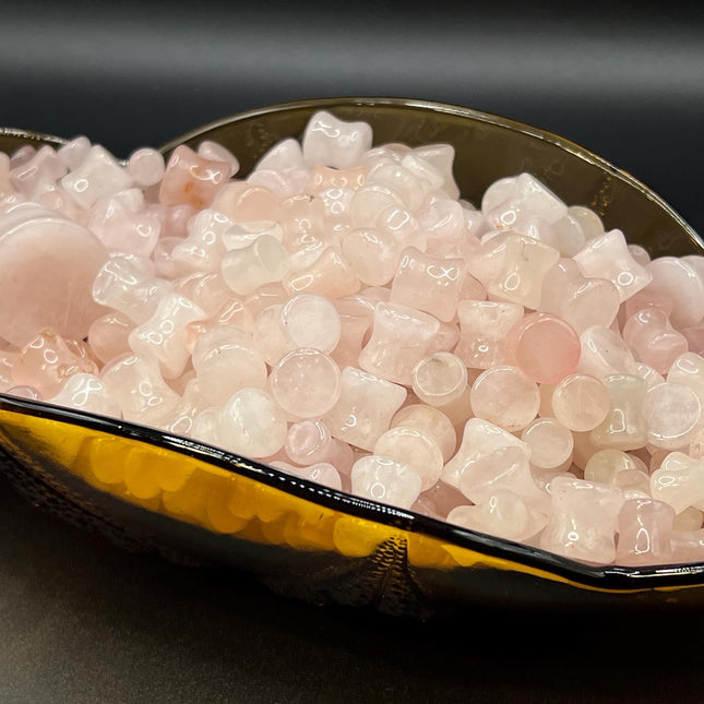 A beautifully polished pair of Rose Quartz Double Flare Plugs rests delicately on a pale surface, their soft pink tones glowing in natural light. Each plug features the gentle, translucent hue that rose quartz is known for—ranging from milky blush to faint petal pink, with subtle natural marbling throughout. The smooth, glossy finish highlights the stone’s calming energy, while the double flare design ensures a secure, comfortable fit for stretched ears.