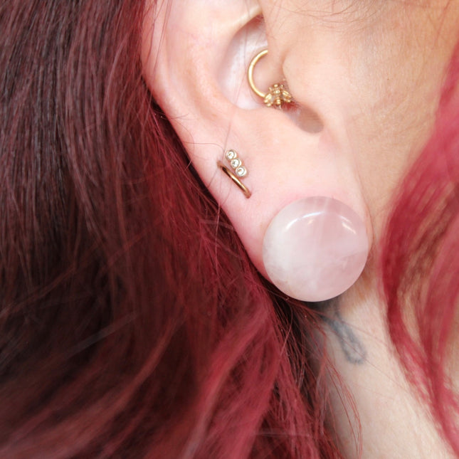 A beautifully polished pair of Rose Quartz Double Flare Plugs rests delicately on a pale surface, their soft pink tones glowing in natural light. Each plug features the gentle, translucent hue that rose quartz is known for—ranging from milky blush to faint petal pink, with subtle natural marbling throughout. The smooth, glossy finish highlights the stone’s calming energy, while the double flare design ensures a secure, comfortable fit for stretched ears.