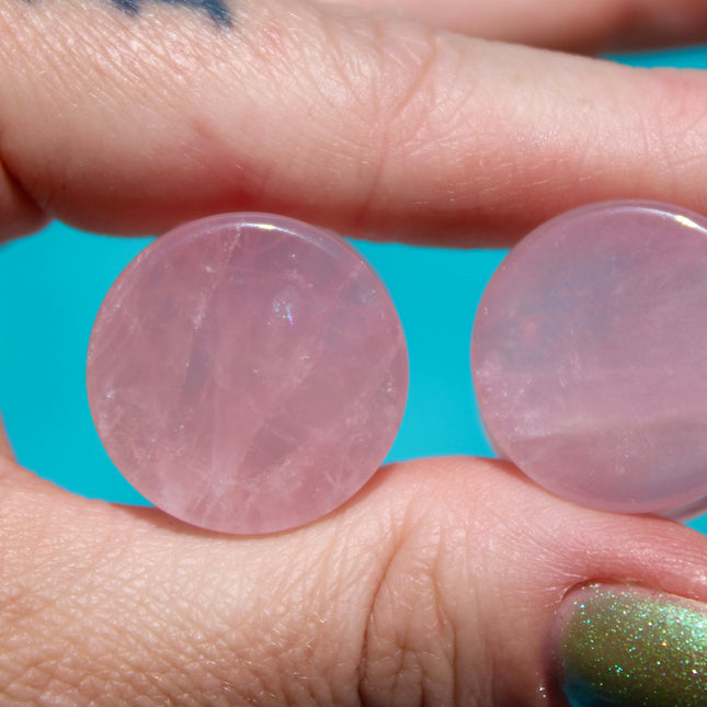 A beautifully polished pair of Rose Quartz Double Flare Plugs rests delicately on a pale surface, their soft pink tones glowing in natural light. Each plug features the gentle, translucent hue that rose quartz is known for—ranging from milky blush to faint petal pink, with subtle natural marbling throughout. The smooth, glossy finish highlights the stone’s calming energy, while the double flare design ensures a secure, comfortable fit for stretched ears.