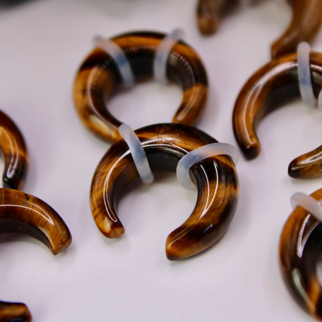 A sleek and powerful Tiger Eye Pincher Gauge rests gracefully against a clean background, glowing with warm golden and brown tones. The curved, crescent shape of the pincher showcases the stone’s natural chatoyancy—those silky, light-catching bands that seem to move as the piece turns. Swirls of honey gold, rich amber, and deep espresso create a striking contrast that’s both earthy and luxurious.