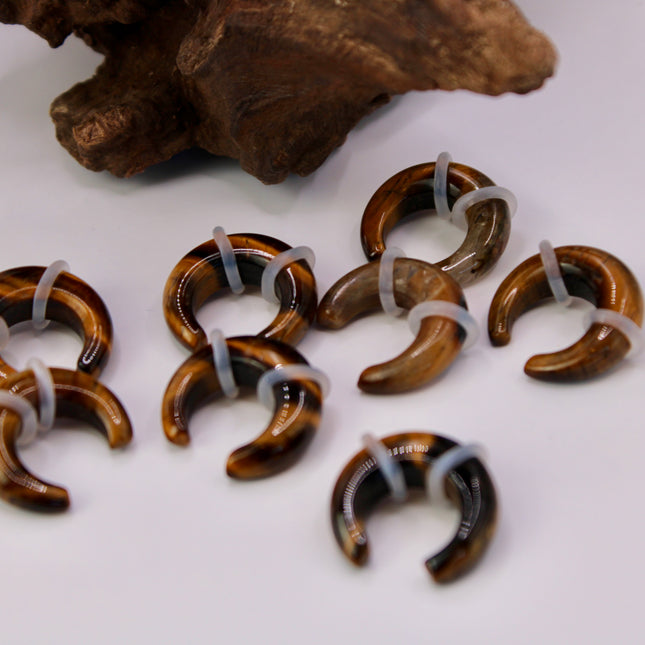 A sleek and powerful Tiger Eye Pincher Gauge rests gracefully against a clean background, glowing with warm golden and brown tones. The curved, crescent shape of the pincher showcases the stone’s natural chatoyancy—those silky, light-catching bands that seem to move as the piece turns. Swirls of honey gold, rich amber, and deep espresso create a striking contrast that’s both earthy and luxurious.