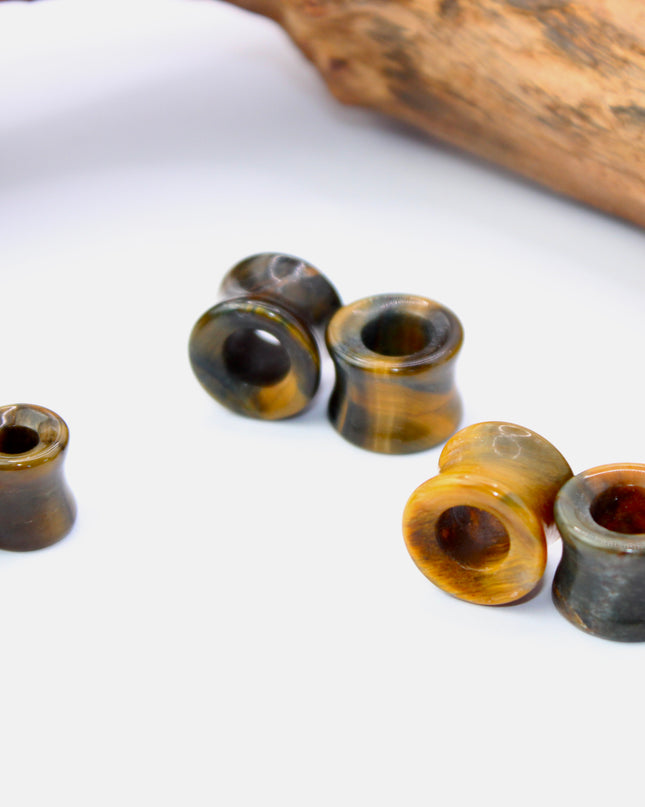 A striking pair of Tiger Eye tunnel plugs, precision-carved and polished to a smooth, glossy finish. These double flare tunnels feature the signature golden-brown hues and shimmering chatoyancy that Tiger Eye is known for, with each piece showcasing unique natural banding and depth. The hollow center adds a bold visual dimension while keeping the plugs lightweight and breathable for stretched ears. 