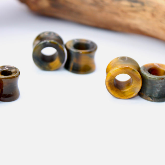 A striking pair of Tiger Eye tunnel plugs, precision-carved and polished to a smooth, glossy finish. These double flare tunnels feature the signature golden-brown hues and shimmering chatoyancy that Tiger Eye is known for, with each piece showcasing unique natural banding and depth. The hollow center adds a bold visual dimension while keeping the plugs lightweight and breathable for stretched ears. 