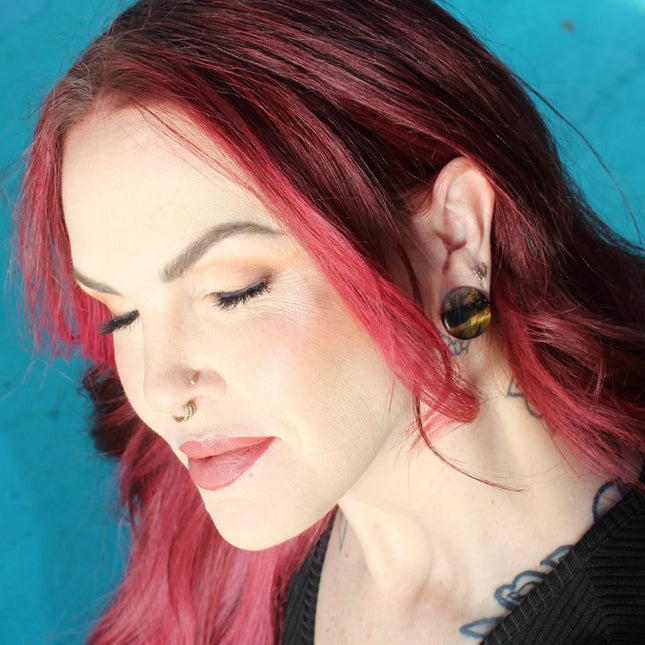 A sleek pair of Tiger Eye Double Flare Stone Plugs rests on a natural-toned backdrop, catching the light with their signature golden-brown shimmer. The polished surface reveals mesmerizing bands of gold, bronze, and deep brown—creating a silky, chatoyant effect that shifts as you move. Each plug is expertly shaped with a subtle double flare for secure, comfortable wear. The rich, earthy tones and natural luster make these plugs both grounding and bold—perfect for those seeking a balance of strength and sty