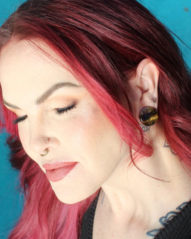 A sleek pair of Tiger Eye Double Flare Stone Plugs rests on a natural-toned backdrop, catching the light with their signature golden-brown shimmer. The polished surface reveals mesmerizing bands of gold, bronze, and deep brown—creating a silky, chatoyant effect that shifts as you move. Each plug is expertly shaped with a subtle double flare for secure, comfortable wear. The rich, earthy tones and natural luster make these plugs both grounding and bold—perfect for those seeking a balance of strength and sty