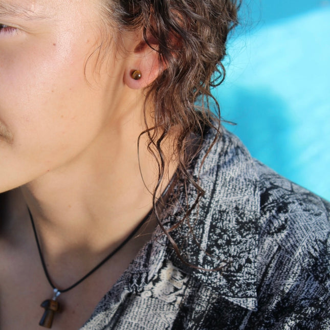 A sleek pair of Tiger Eye Double Flare Stone Plugs rests on a natural-toned backdrop, catching the light with their signature golden-brown shimmer. The polished surface reveals mesmerizing bands of gold, bronze, and deep brown—creating a silky, chatoyant effect that shifts as you move. Each plug is expertly shaped with a subtle double flare for secure, comfortable wear. The rich, earthy tones and natural luster make these plugs both grounding and bold—perfect for those seeking a balance of strength and sty