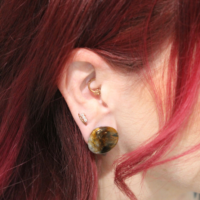 A sleek pair of Tiger Eye Double Flare Stone Plugs rests on a natural-toned backdrop, catching the light with their signature golden-brown shimmer. The polished surface reveals mesmerizing bands of gold, bronze, and deep brown—creating a silky, chatoyant effect that shifts as you move. Each plug is expertly shaped with a subtle double flare for secure, comfortable wear. The rich, earthy tones and natural luster make these plugs both grounding and bold—perfect for those seeking a balance of strength and sty