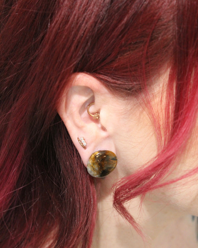 A sleek pair of Tiger Eye Double Flare Stone Plugs rests on a natural-toned backdrop, catching the light with their signature golden-brown shimmer. The polished surface reveals mesmerizing bands of gold, bronze, and deep brown—creating a silky, chatoyant effect that shifts as you move. Each plug is expertly shaped with a subtle double flare for secure, comfortable wear. The rich, earthy tones and natural luster make these plugs both grounding and bold—perfect for those seeking a balance of strength and sty