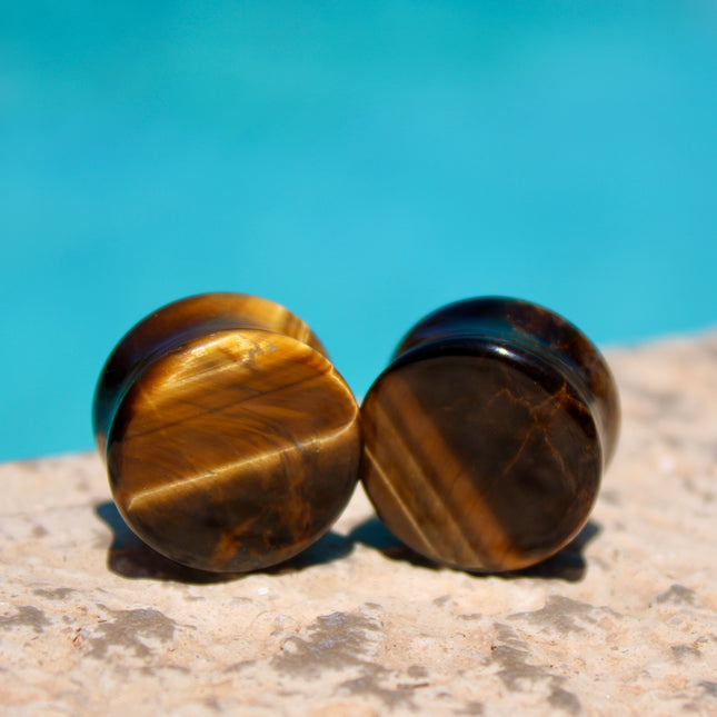 A sleek pair of Tiger Eye Double Flare Stone Plugs rests on a natural-toned backdrop, catching the light with their signature golden-brown shimmer. The polished surface reveals mesmerizing bands of gold, bronze, and deep brown—creating a silky, chatoyant effect that shifts as you move. Each plug is expertly shaped with a subtle double flare for secure, comfortable wear. The rich, earthy tones and natural luster make these plugs both grounding and bold—perfect for those seeking a balance of strength and sty