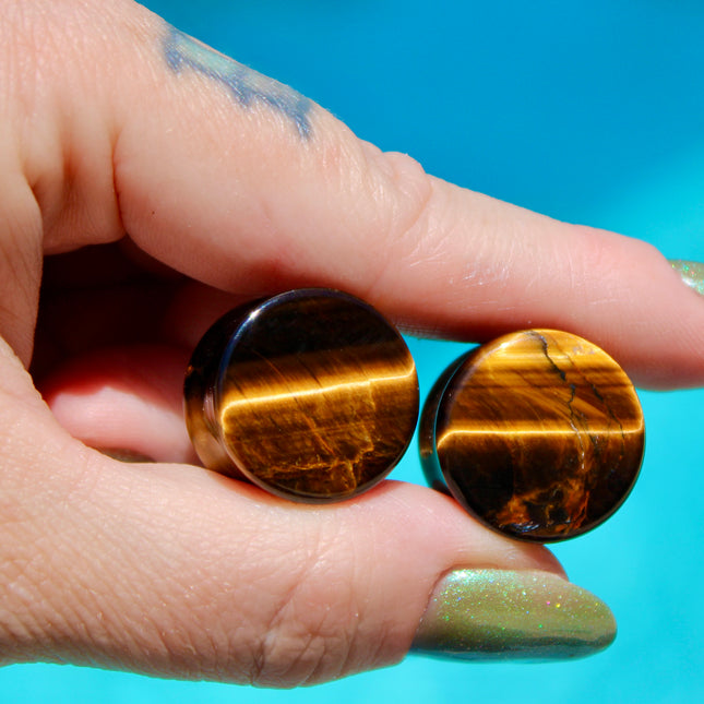 A sleek pair of Tiger Eye Double Flare Stone Plugs rests on a natural-toned backdrop, catching the light with their signature golden-brown shimmer. The polished surface reveals mesmerizing bands of gold, bronze, and deep brown—creating a silky, chatoyant effect that shifts as you move. Each plug is expertly shaped with a subtle double flare for secure, comfortable wear. The rich, earthy tones and natural luster make these plugs both grounding and bold—perfect for those seeking a balance of strength and sty