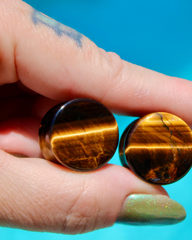 
A sleek pair of Tiger Eye Double Flare Stone Plugs rests on a natural-toned backdrop, catching the light with their signature golden-brown shimmer. The polished surface reveals mesmerizing bands of gold, bronze, and deep brown—creating a silky, chatoyant effect that shifts as you move. Each plug is expertly shaped with a subtle double flare for secure, comfortable wear. The rich, earthy tones and natural luster make these plugs both grounding and bold—perfect for those seeking a balance of strength and sty