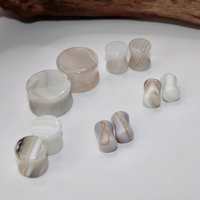 A serene pair of White Line Agate Plugs rests on a soft, neutral background, showcasing their natural, minimalist beauty. Each plug features delicate layers of white, cream, and translucent tones, with subtle linear banding unique to every piece. The polished finish enhances the stone’s soft sheen and calming aesthetic. 