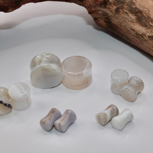 A serene pair of White Line Agate Plugs rests on a soft, neutral background, showcasing their natural, minimalist beauty. Each plug features delicate layers of white, cream, and translucent tones, with subtle linear banding unique to every piece. The polished finish enhances the stone’s soft sheen and calming aesthetic. 