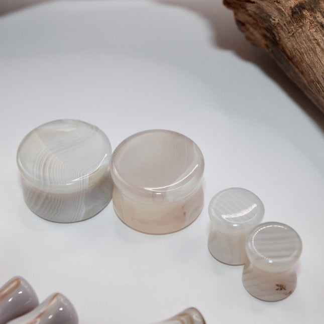 A serene pair of White Line Agate Plugs rests on a soft, neutral background, showcasing their natural, minimalist beauty. Each plug features delicate layers of white, cream, and translucent tones, with subtle linear banding unique to every piece. The polished finish enhances the stone’s soft sheen and calming aesthetic. 
