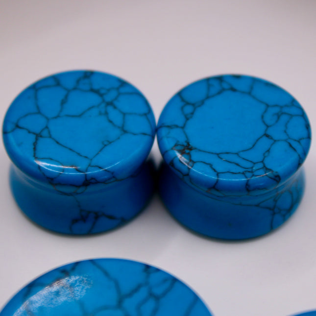 A vibrant pair of Blue Howlite Stone Plugs is shown resting on a soft beige surface. The plugs are round, polished to a high shine, and display a brilliant turquoise-blue color. Each plug is marked with distinctive dark gray to black veining in a web-like pattern—natural to howlite that has been dyed blue to resemble turquoise. The smooth, glossy finish enhances the contrast between the bright blue background and the delicate marbling. The lighting gently reflects off the curved surfaces, emphasizing their 
