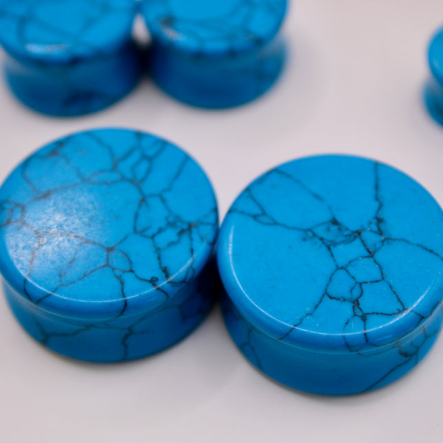 A vibrant pair of Blue Howlite Stone Plugs is shown resting on a soft beige surface. The plugs are round, polished to a high shine, and display a brilliant turquoise-blue color. Each plug is marked with distinctive dark gray to black veining in a web-like pattern—natural to howlite that has been dyed blue to resemble turquoise. The smooth, glossy finish enhances the contrast between the bright blue background and the delicate marbling. The lighting gently reflects off the curved surfaces, emphasizing their