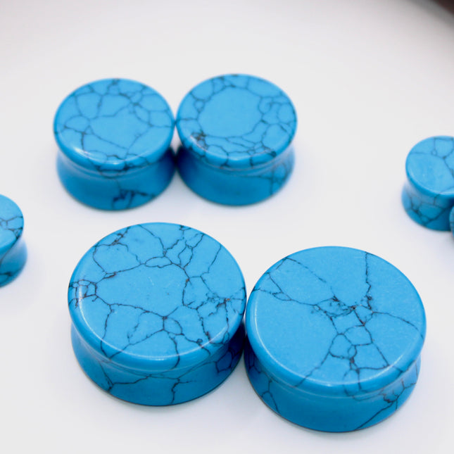 A vibrant pair of Blue Howlite Stone Plugs is shown resting on a soft beige surface. The plugs are round, polished to a high shine, and display a brilliant turquoise-blue color. Each plug is marked with distinctive dark gray to black veining in a web-like pattern—natural to howlite that has been dyed blue to resemble turquoise. The smooth, glossy finish enhances the contrast between the bright blue background and the delicate marbling. The lighting gently reflects off the curved surfaces, emphasizing their