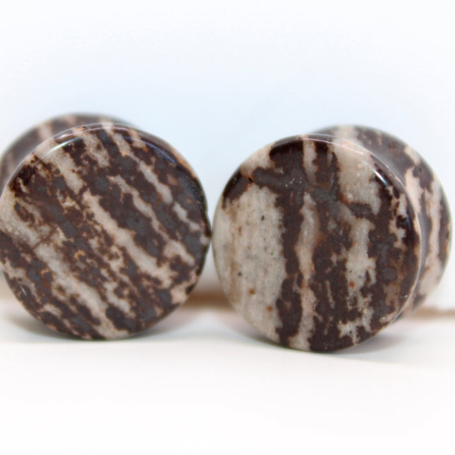 A striking pair of Natural Hematite Brown Double Flared Plugs rests against a soft neutral background, their deep, earthy tones catching the light with a smooth metallic sheen. Each plug is truly one of a kind, showcasing a rich blend of dark brown, bronze, and gunmetal hues with subtle shimmer—hallmarks of natural hematite with unique mineral variations. The double flare design ensures a snug and comfortable fit, while the polished surface adds a sleek, reflective quality. 