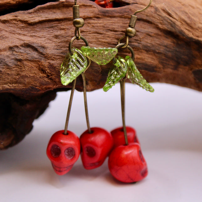 A close-up of a pair of striking Gothic earrings featuring glossy, deep-red cherry charms hanging from small silver skulls. The cherries appear juicy and vibrant, with a subtle dark shine, while the skulls add a bold, edgy contrast. The earrings are displayed against a black velvet background, enhancing their rich colors and alt-aesthetic vibe. Perfect for alternative, punk, or gothic fashion lovers who enjoy mixing sweetness with a hint of rebellion.