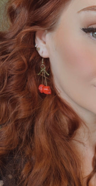 A close-up of a pair of striking Gothic earrings featuring glossy, deep-red cherry charms hanging from small silver skulls. The cherries appear juicy and vibrant, with a subtle dark shine, while the skulls add a bold, edgy contrast. The earrings are displayed against a black velvet background, enhancing their rich colors and alt-aesthetic vibe. Perfect for alternative, punk, or gothic fashion lovers who enjoy mixing sweetness with a hint of rebellion.