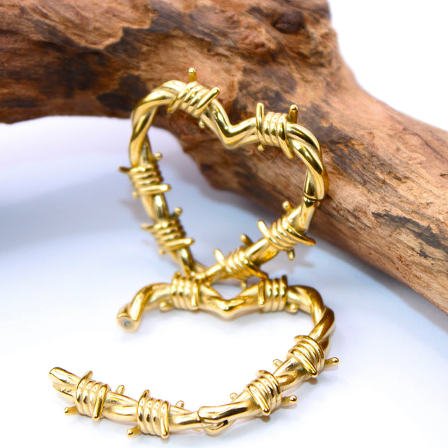 A striking pair of heart-shaped hangers designed for stretched ears, crafted in a shiny gold-tone metal with detailed barbed wire accents wrapping around the heart outline. The edgy design blends romance with rebellion, featuring smooth curves contrasted by sharp, sculpted wire barbs for a bold, punk-inspired aesthetic. Made to hang comfortably in stretched lobes, these hangers are lightweight yet visually impactful—perfect for expressing individuality with attitude and style.