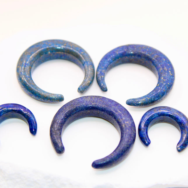 A striking Genuine Lapis Lazuli Stone Septum Gauge rests gently against a neutral-toned surface. The deep royal blue of the stone is rich and captivating, marbled with subtle veins of white calcite and scattered golden flecks of pyrite—classic hallmarks of authentic lapis. Expertly carved and polished into a smooth, curved shape for septum wear, the piece blends bold elegance with earthy mystique. Its surface gleams under soft lighting, showcasing the depth and natural beauty of the stone