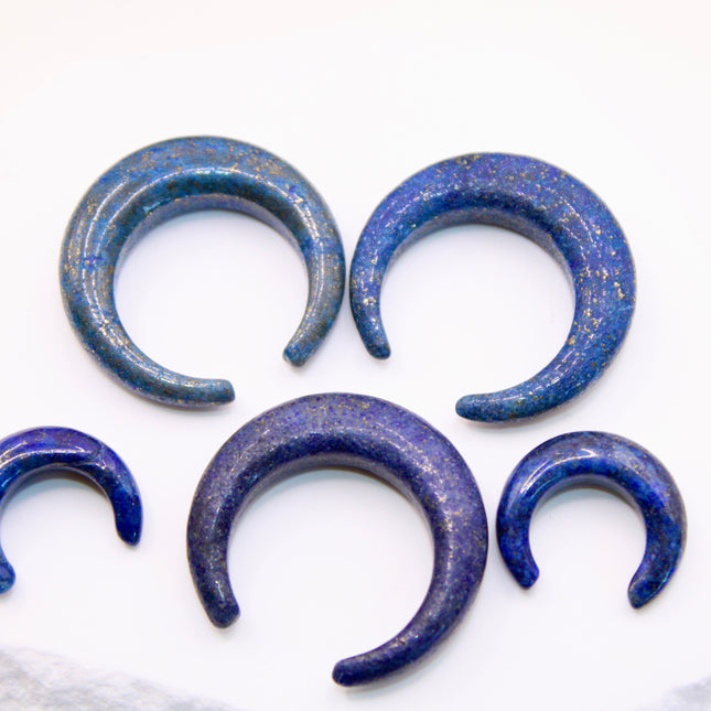 A striking Genuine Lapis Lazuli Stone Septum Gauge rests gently against a neutral-toned surface. The deep royal blue of the stone is rich and captivating, marbled with subtle veins of white calcite and scattered golden flecks of pyrite—classic hallmarks of authentic lapis. Expertly carved and polished into a smooth, curved shape for septum wear, the piece blends bold elegance with earthy mystique. Its surface gleams under soft lighting, showcasing the depth and natural beauty of the stone