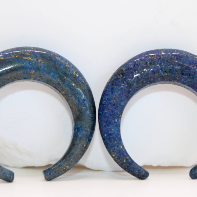 A striking Genuine Lapis Lazuli Stone Septum Gauge rests gently against a neutral-toned surface. The deep royal blue of the stone is rich and captivating, marbled with subtle veins of white calcite and scattered golden flecks of pyrite—classic hallmarks of authentic lapis. Expertly carved and polished into a smooth, curved shape for septum wear, the piece blends bold elegance with earthy mystique. Its surface gleams under soft lighting, showcasing the depth and natural beauty of the stone