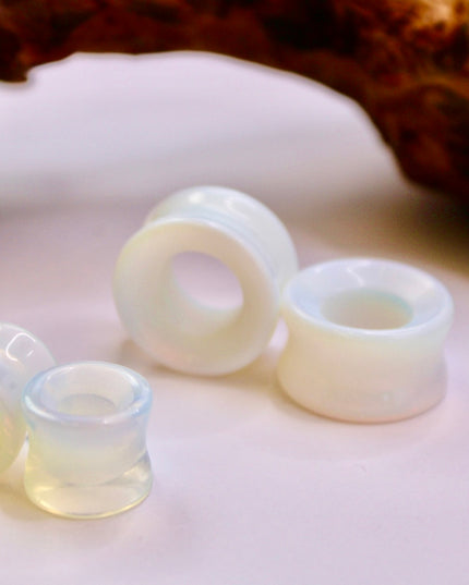 A radiant pair of Opalite Tunnel Rainbow Plugs glows softly against a neutral background, their translucent bodies catching and bending light with a subtle, ethereal rainbow effect. The opalite’s milky, iridescent surface shifts between soft blues, purples, and warm golden tones as the light moves, creating a magical, otherworldly glow. Designed as tunnels, these plugs are lightweight and comfortable, with a smooth double flare for a secure fit. 