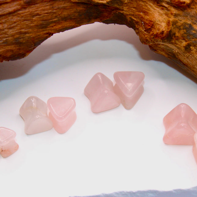 A graceful pair of Rose Quartz Triangle Double Flared Plugs lies elegantly on a soft, neutral surface. Each plug is carved into a bold triangle shape with gently rounded edges, offering a unique geometric twist on classic plug designs. The soft pink hue of the rose quartz glows subtly in the light, with natural marbling and translucent tones giving each piece its own delicate character. Smoothly polished for comfort and shaped with a double flare for a secure fit, these plugs blend crystal calm with modern 