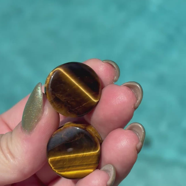 A sleek pair of Tiger Eye Double Flare Stone Plugs rests on a natural-toned backdrop, catching the light with their signature golden-brown shimmer. The polished surface reveals mesmerizing bands of gold, bronze, and deep brown—creating a silky, chatoyant effect that shifts as you move. Each plug is expertly shaped with a subtle double flare for secure, comfortable wear. The rich, earthy tones and natural luster make these plugs both grounding and bold—perfect for those seeking a balance of strength and sty