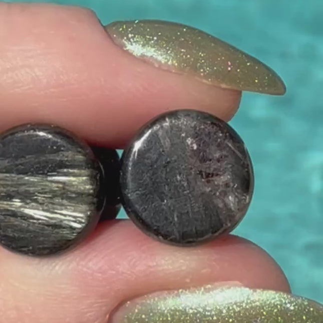A stunning pair of Astrophyllite FLASH Double Flare Plugs rests against a dark, textured backdrop, their surfaces gleaming with bursts of golden-bronze metallic flash. Each plug is crafted from genuine astrophyllite, a rare and complex stone known for its radiant, starburst-like inclusions that shimmer brilliantly under light. The deep black and charcoal base is layered with reflective veins and flashes of copper, gold, and occasionally blue—giving these plugs a cosmic, otherworldly appearance.