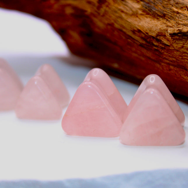 A graceful pair of Rose Quartz Triangle Double Flared Plugs lies elegantly on a soft, neutral surface. Each plug is carved into a bold triangle shape with gently rounded edges, offering a unique geometric twist on classic plug designs. The soft pink hue of the rose quartz glows subtly in the light, with natural marbling and translucent tones giving each piece its own delicate character. Smoothly polished for comfort and shaped with a double flare for a secure fit, these plugs blend crystal calm with modern 