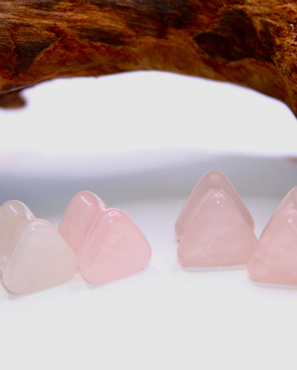 A graceful pair of Rose Quartz Triangle Double Flared Plugs lies elegantly on a soft, neutral surface. Each plug is carved into a bold triangle shape with gently rounded edges, offering a unique geometric twist on classic plug designs. The soft pink hue of the rose quartz glows subtly in the light, with natural marbling and translucent tones giving each piece its own delicate character. Smoothly polished for comfort and shaped with a double flare for a secure fit, these plugs blend crystal calm with modern 