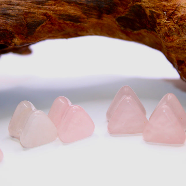 A graceful pair of Rose Quartz Triangle Double Flared Plugs lies elegantly on a soft, neutral surface. Each plug is carved into a bold triangle shape with gently rounded edges, offering a unique geometric twist on classic plug designs. The soft pink hue of the rose quartz glows subtly in the light, with natural marbling and translucent tones giving each piece its own delicate character. Smoothly polished for comfort and shaped with a double flare for a secure fit, these plugs blend crystal calm with modern 