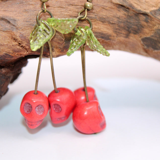 A close-up of a pair of striking Gothic earrings featuring glossy, deep-red cherry charms hanging from small silver skulls. The cherries appear juicy and vibrant, with a subtle dark shine, while the skulls add a bold, edgy contrast. The earrings are displayed against a black velvet background, enhancing their rich colors and alt-aesthetic vibe. Perfect for alternative, punk, or gothic fashion lovers who enjoy mixing sweetness with a hint of rebellion.