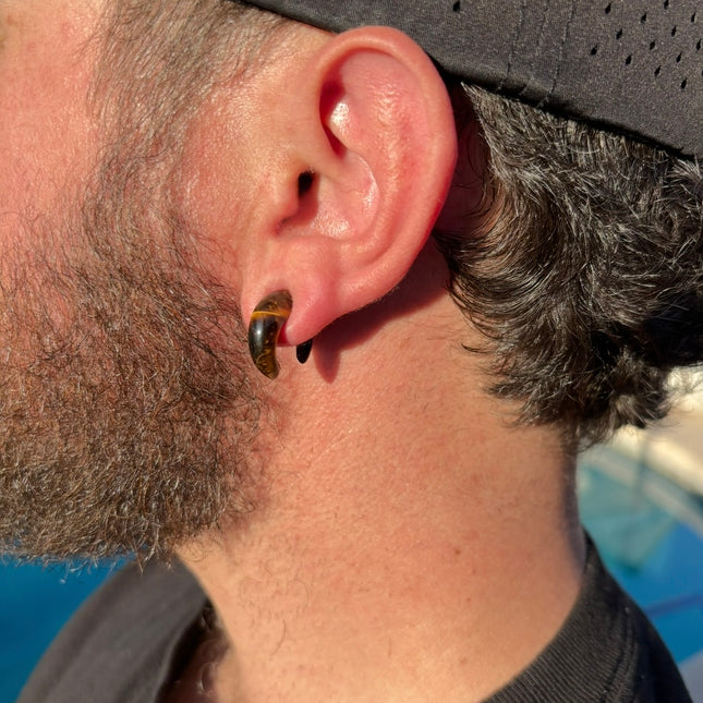 A sleek and powerful Tiger Eye Pincher Gauge rests gracefully against a clean background, glowing with warm golden and brown tones. The curved, crescent shape of the pincher showcases the stone’s natural chatoyancy—those silky, light-catching bands that seem to move as the piece turns. Swirls of honey gold, rich amber, and deep espresso create a striking contrast that’s both earthy and luxurious.