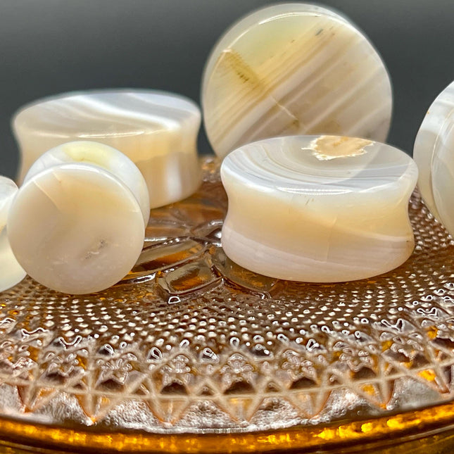 A serene pair of White Line Agate Plugs rests on a soft, neutral background, showcasing their natural, minimalist beauty. Each plug features delicate layers of white, cream, and translucent tones, with subtle linear banding unique to every piece. The polished finish enhances the stone’s soft sheen and calming aesthetic. 