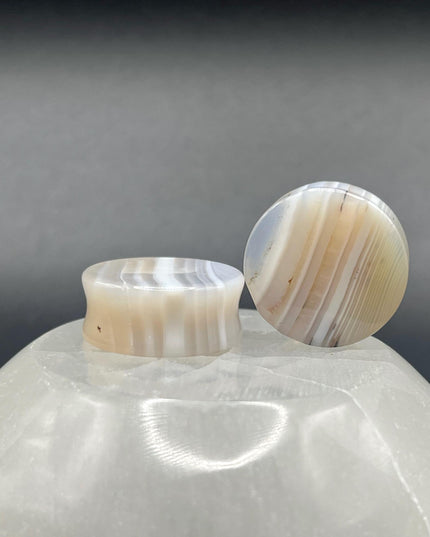 A serene pair of White Line Agate Plugs rests on a soft, neutral background, showcasing their natural, minimalist beauty. Each plug features delicate layers of white, cream, and translucent tones, with subtle linear banding unique to every piece. The polished finish enhances the stone’s soft sheen and calming aesthetic. 