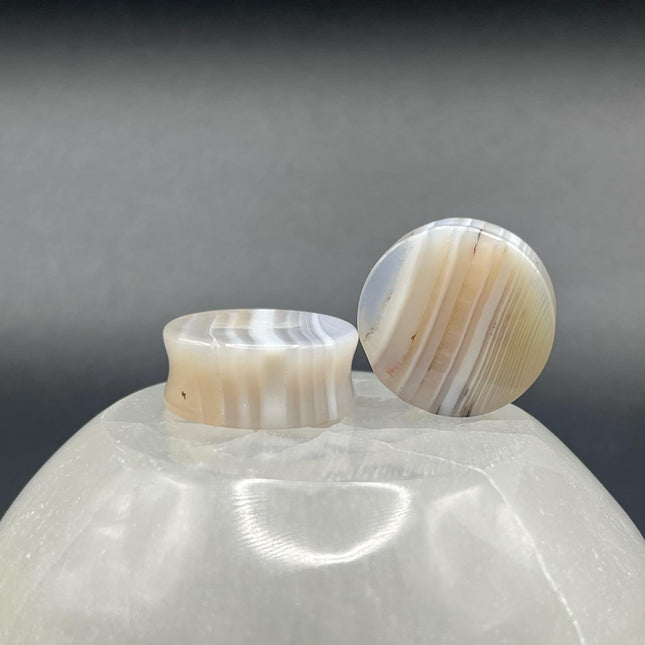 A serene pair of White Line Agate Plugs rests on a soft, neutral background, showcasing their natural, minimalist beauty. Each plug features delicate layers of white, cream, and translucent tones, with subtle linear banding unique to every piece. The polished finish enhances the stone’s soft sheen and calming aesthetic.
