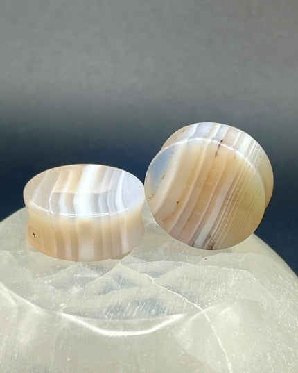 A serene pair of White Line Agate Plugs rests on a soft, neutral background, showcasing their natural, minimalist beauty. Each plug features delicate layers of white, cream, and translucent tones, with subtle linear banding unique to every piece. The polished finish enhances the stone’s soft sheen and calming aesthetic. 