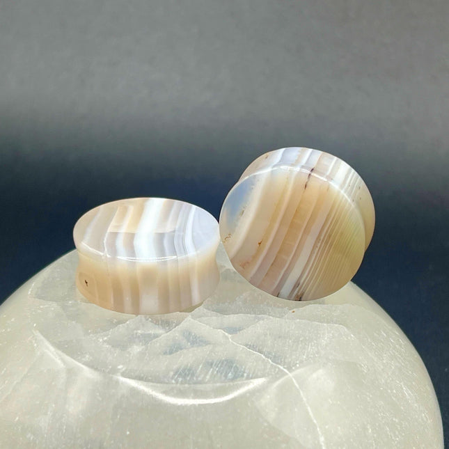 A serene pair of White Line Agate Plugs rests on a soft, neutral background, showcasing their natural, minimalist beauty. Each plug features delicate layers of white, cream, and translucent tones, with subtle linear banding unique to every piece. The polished finish enhances the stone’s soft sheen and calming aesthetic. 